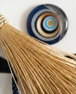 Μάκρο φωτογραφία που εστιάζει στη χρυσή φούντα, το μεταλλικό δέσιμο, το navy κορδόνι και τους χρυσούς κύκλους στο γυάλινο ματάκι.