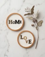 Μίνιμαλ σετ σουβέρ HoMe και LoVE από φελλό τοποθετημένα σε λευκή μαρμάρινη επιφάνεια δίπλα σε ένα αποξηραμένο κλαδί.