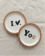 Μίνιμαλ σουβέρ φελλού και πορσελάνης I Love You φωτογραφημένα από πάνω, πάνω σε ύφασμα με λινή υφή.