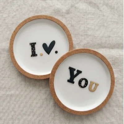 Μίνιμαλ σουβέρ φελλού και πορσελάνης I Love You φωτογραφημένα από πάνω, πάνω σε ύφασμα με λινή υφή.